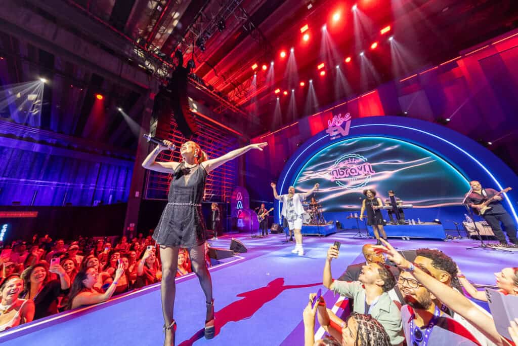 Imagem de show ao vivo com artista e banda em palco iluminado, público animado e cenário moderno.