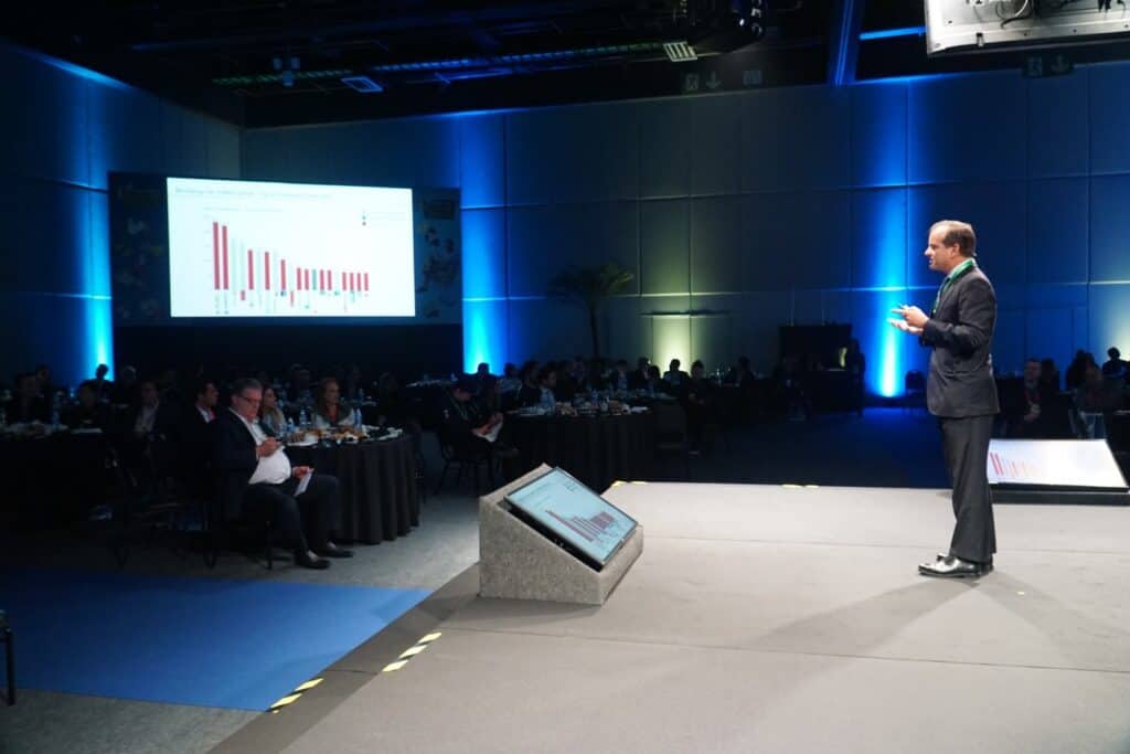 Apresentador em palco durante conferência de negócios com gráfico de barras na tela.